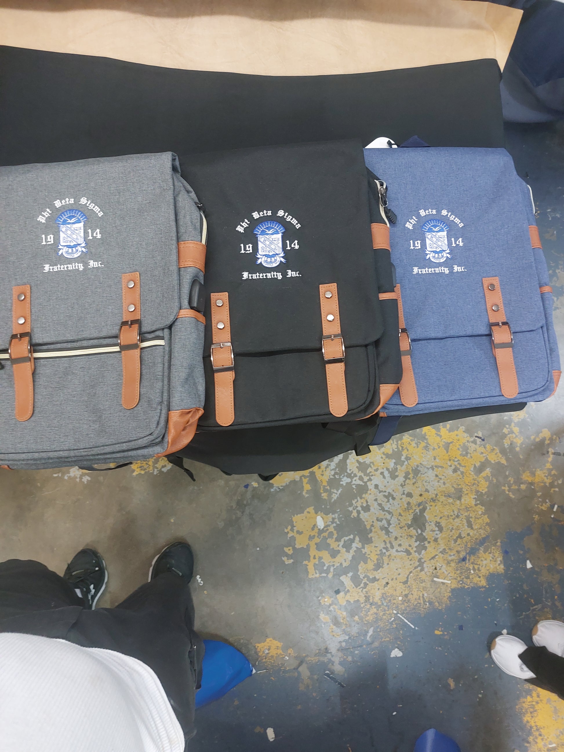 A collection of Phi Beta Sigma backpacks in black and blue colors, featuring leather accents and a logo on the front.