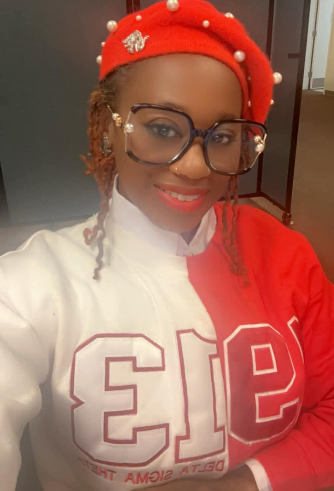 A person wearing a red and white Delta Sigma Theta split sweatshirt, glasses, and a beret with a pom-pom on top.
