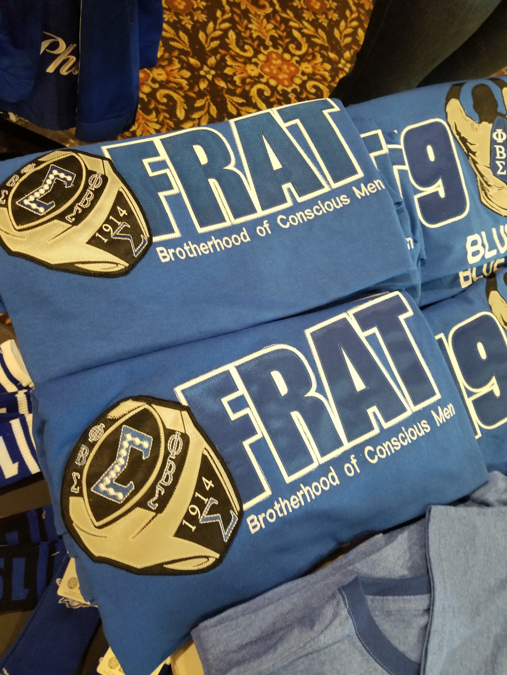 A blue shirt with the text 'FRAT' and the logo of Phi Beta Sigma fraternity. The shirt is displayed in a pile with other similar shirts.