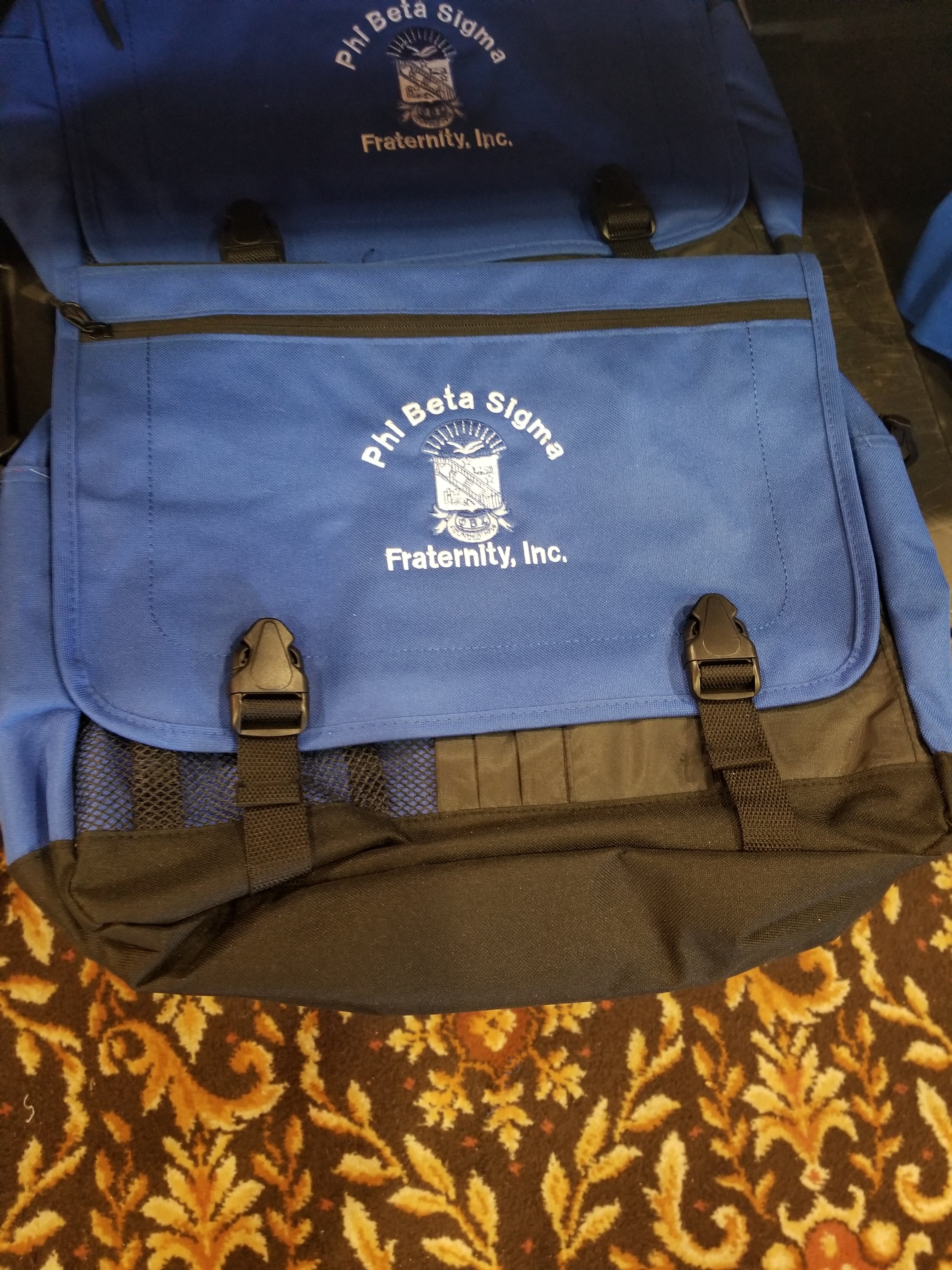 A blue messenger bag with the Phi Beta Sigma fraternity logo on the front. The bag has multiple compartments and is displayed on a patterned surface.