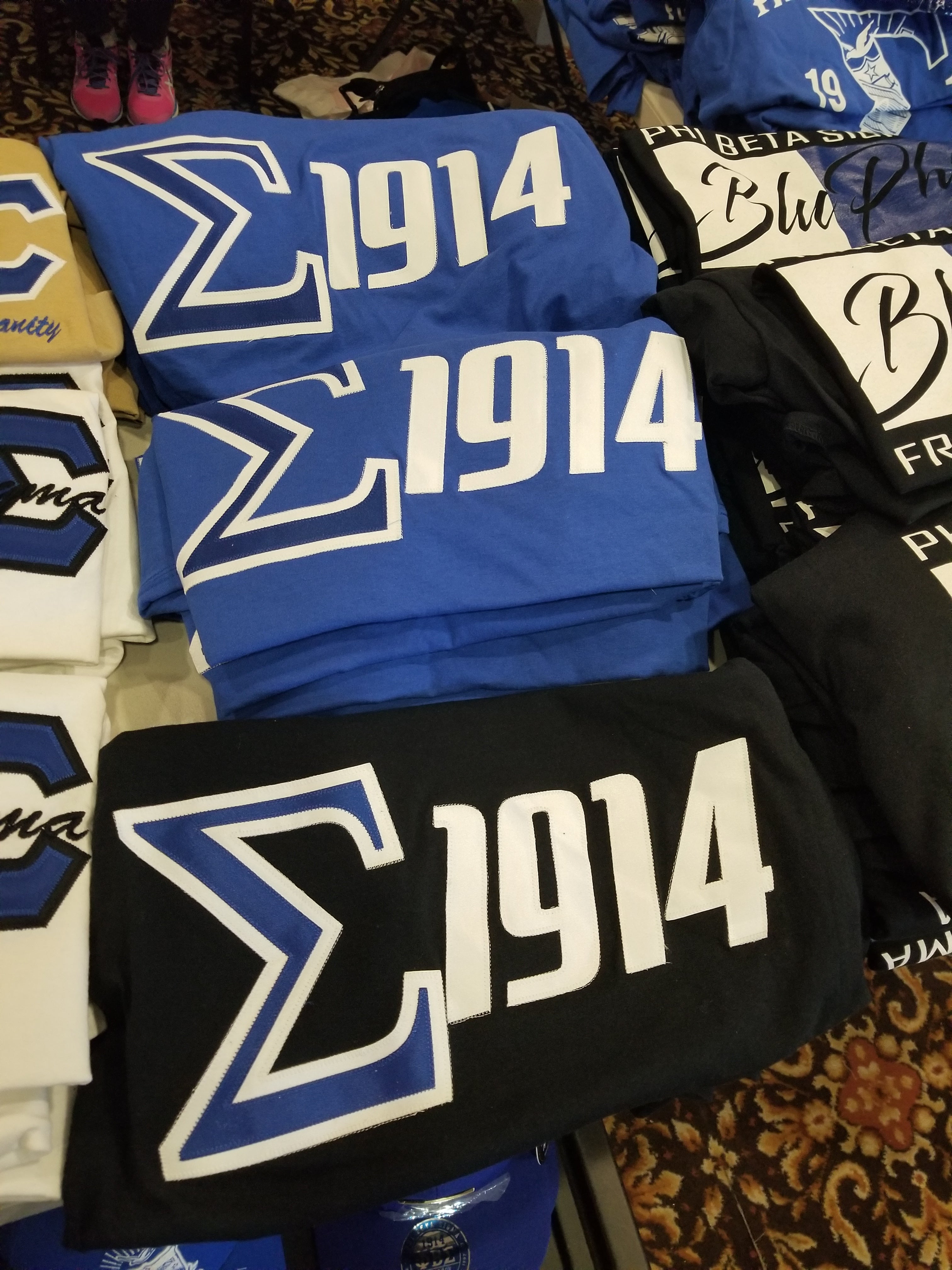 A collection of Phi Beta Sigma shirts displayed, featuring the organization's symbols and the year 1914.