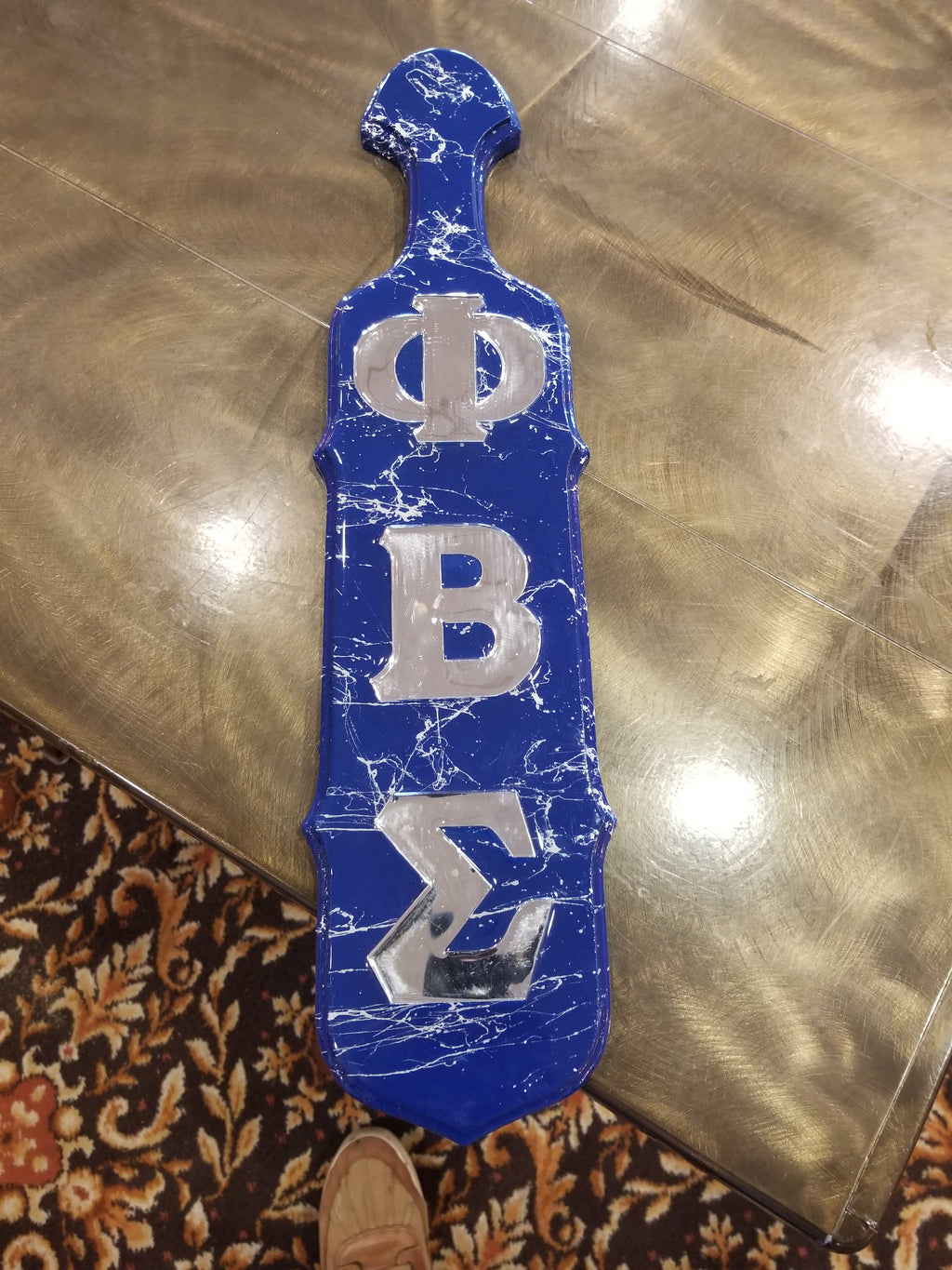 A blue and white resin paddle with the letters 'Zeta' and 'Phi' and the name 'Beta' printed on it, placed on a shiny surface.