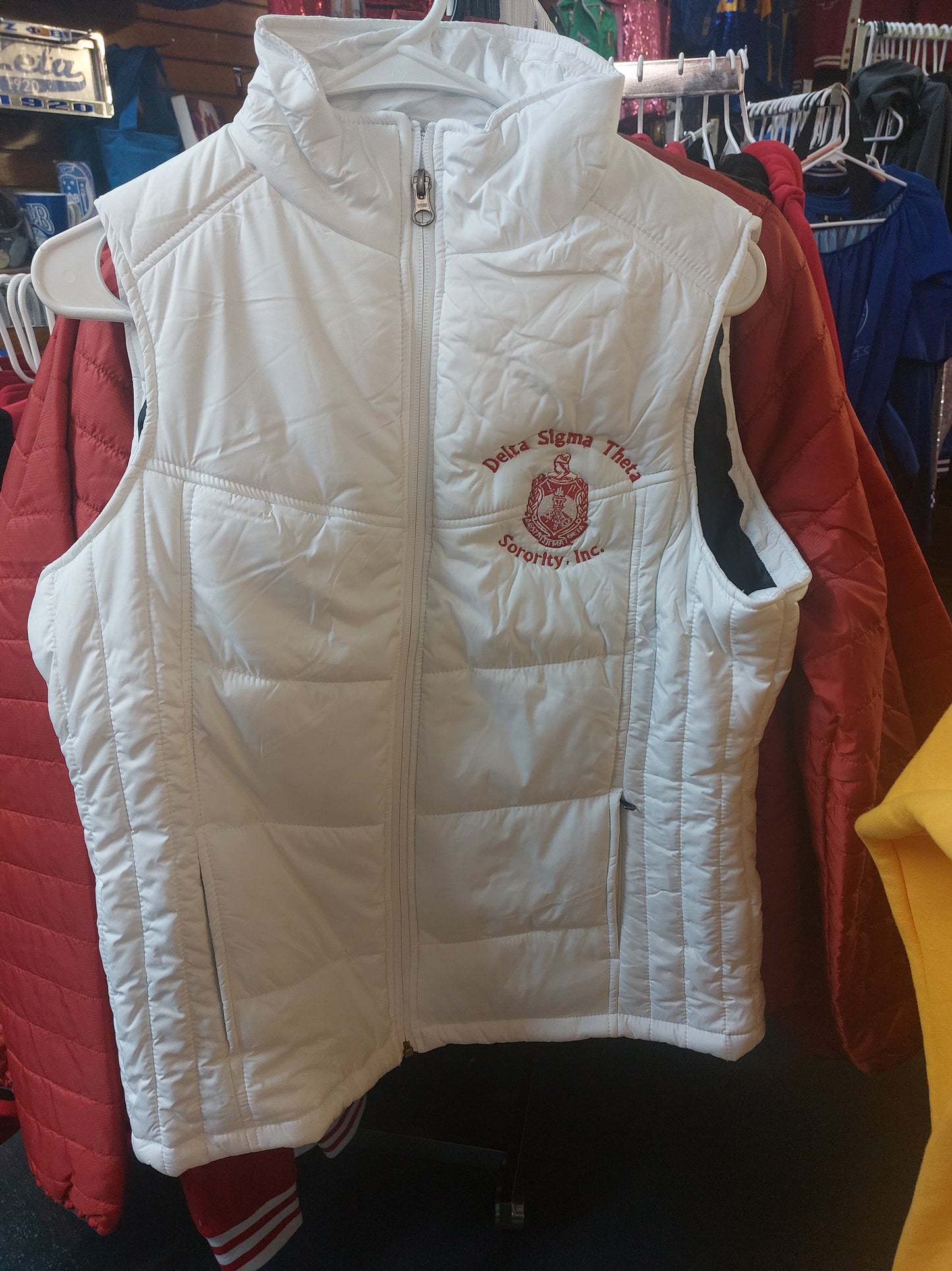 A white Delta Sigma Theta puffer vest with a red and black logo on the front, displayed on a hanger with other clothing items in the background.