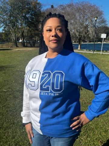 A woman standing outdoors wearing a white and blue split sweatshirt with '1990 Zeta Phi Beta' printed on the front.